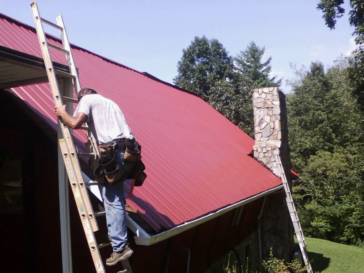 Roofer on a ladder beside a red metal roof and stone chimney — Soffit & Fascia Repair by Roofing Brothers in Monroe Bridge
